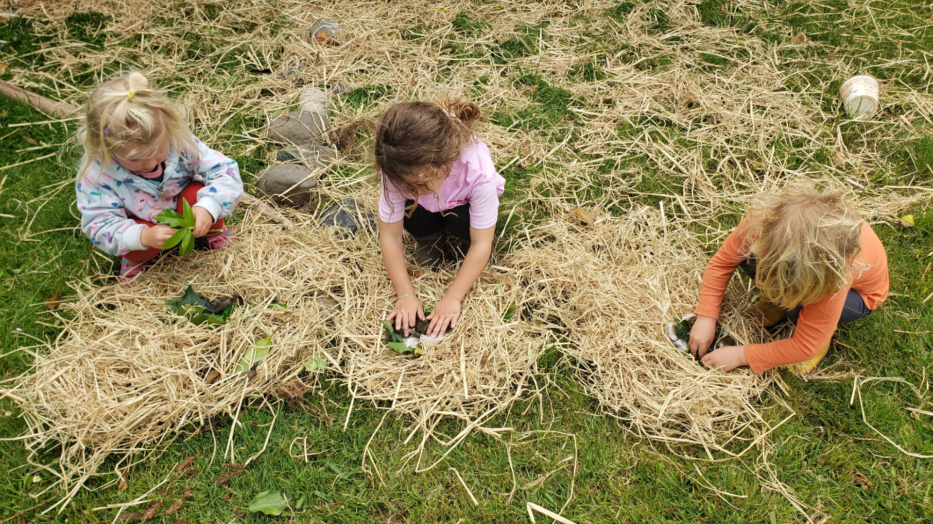 Three children kneeling in straw mulch, planting seedlings by hand